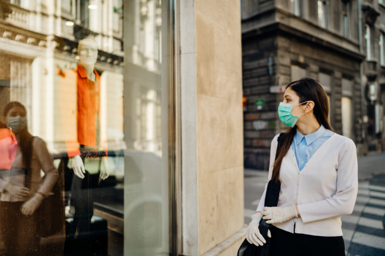 Woman with mask looking at a closed fashion clothes storefront.Clothing shopping during coronavirus outbreak shutdown.COVID-19 quarantine apparel retail store closures.Small business loss concept